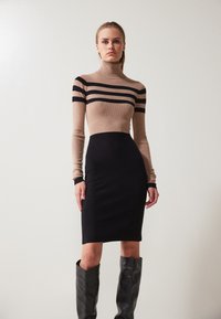 Strickkleid mit einem beigefarbenen Oberteil, das schwarze horizontale Streifen aufweist, und einem engen schwarzen Rock. Lange Ärmel mit kontrastierenden Bündchen; kniehohe schwarze Stiefel.