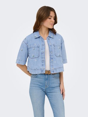 Giovane donna che indossa una giacca di jeans corta azzurro chiaro con ricamo di fiori bianchi sopra una maglietta bianca e jeans blu, in piedi con le mani dietro la schiena.