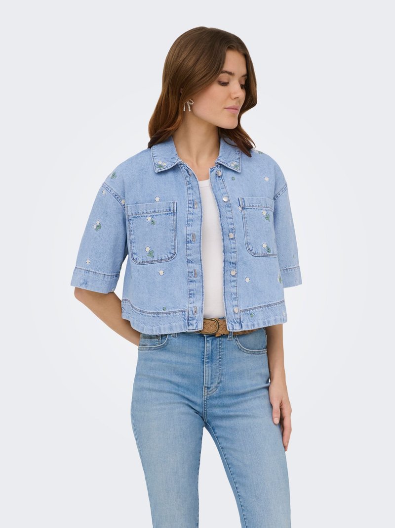 Jeune femme portant une veste en jean courte bleu clair avec une broderie de fleurs blanches sur un top blanc et un jean bleu, debout les mains derrière le dos.