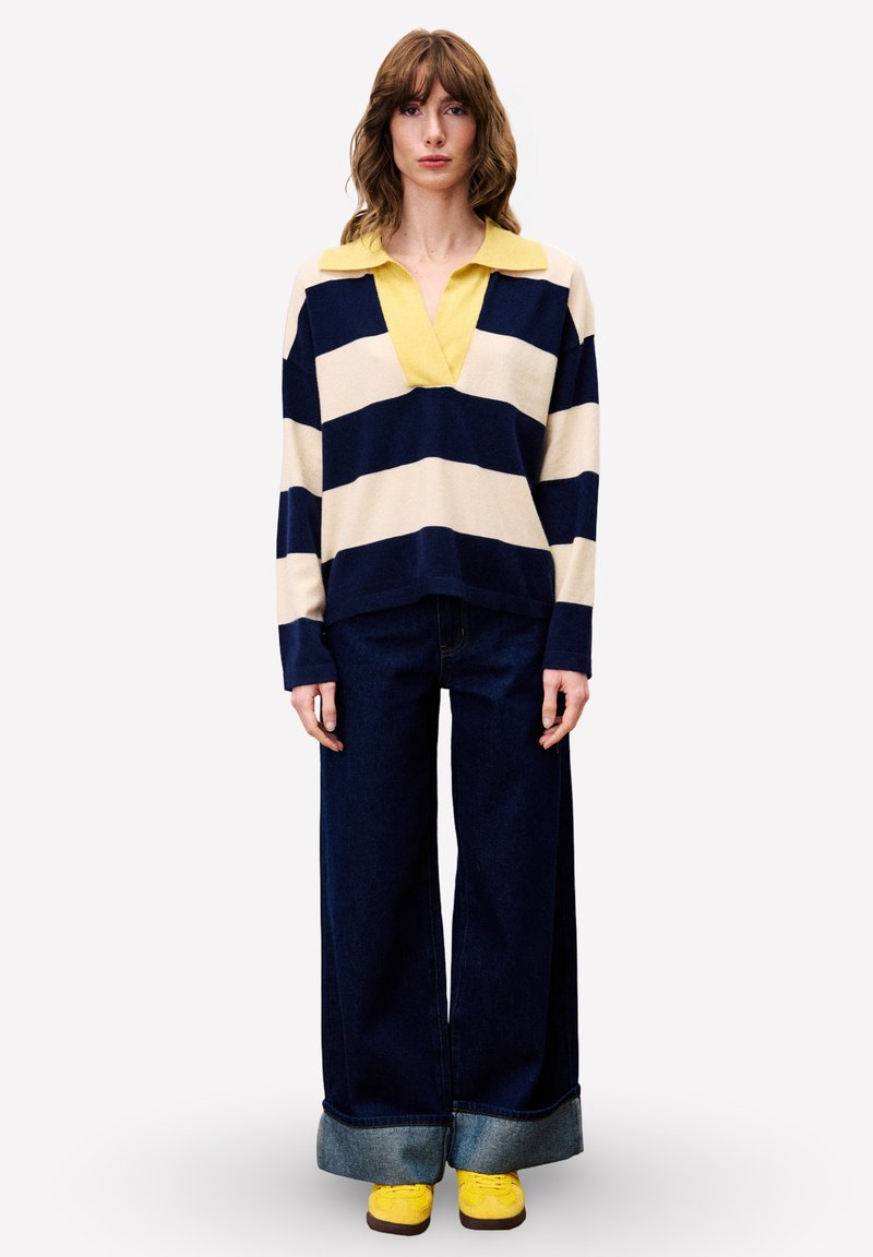 Gestreifter, gestrickter Pullover in Creme und Marineblau mit einem gelben Kragen; kombiniert mit weit geschnittenen, dunkelblauen Jeans und gelben Sneakers.