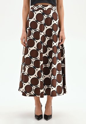 Femme portant une jupe midi marron et blanche à motif géométrique circulaire, un haut noir et des talons aiguilles noirs, debout sur un fond blanc.