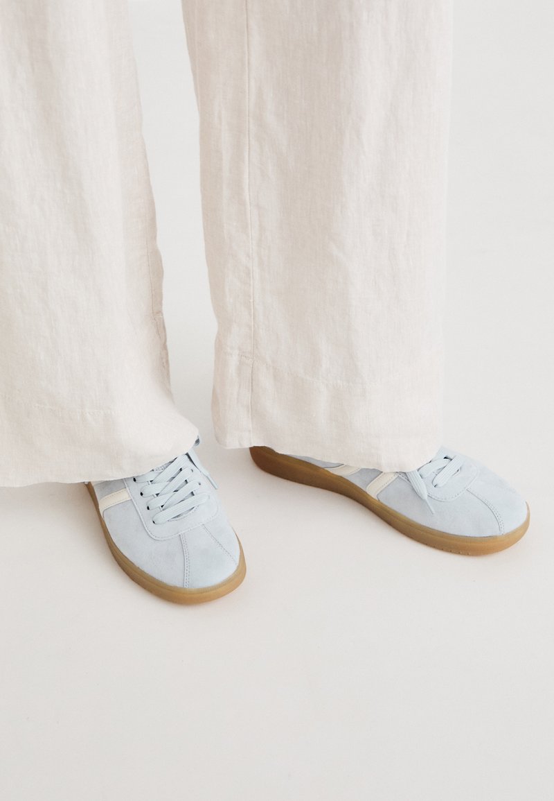 Light blue suede sneakers with white accents, round toe, and flat gum sole, paired with cream-colored, wide linen pants.