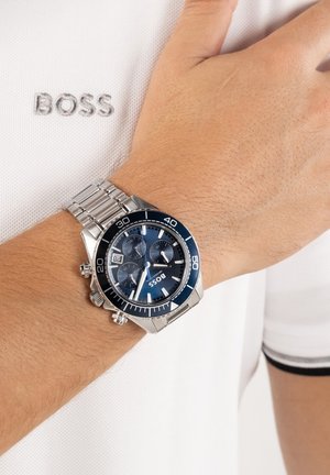 Montre chronographe en métal argenté avec cadran bleu au poignet, portée sur un polo blanc avec "BOSS" brodé sur la poitrine.