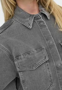 Grå denimskjorta med spetsig krage, två framfickor, texturerad tyg och synliga sömsdetaljer.