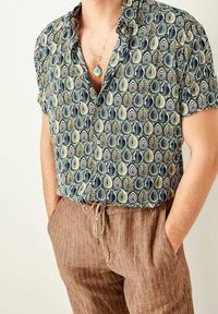 Kurzarmhemd in Grün, Marineblau und Creme mit einem Blatt- und Tränenmuster; kombiniert mit braunen gestreiften Jogginghosen mit Kordelzug.