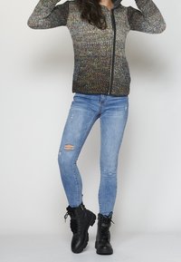 Multikleurige ritstrui met een gestructureerd breipatroon, gecombineerd met lichtblauwe distressed skinny jeans en zwarte veterschoenen tot de enkel.