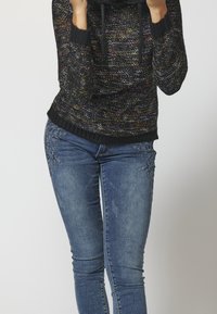 Černý pletený svetr s texturovaným vzorem a kapucí, doplněný modrými džíny s květinovou výšivkou na stehnech a přiléhavým střihem.