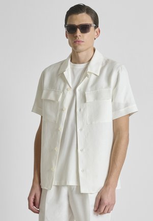 Hombre con gafas de sol oscuras, camisa blanca de manga corta con bolsillos en el pecho, camiseta blanca y pantalones blancos sobre un fondo liso.