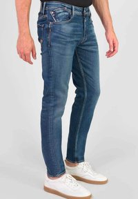 Slim-fit blauwe denim jeans met een lichte vervaging, vijf zakken en contrasterende stiksel, gecombineerd met witte sneakers met een tan kleurige zool.