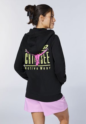 Woman with hair clip wearing a black hoodie with neon pink graphic and yellow text, paired with light purple shorts, facing sideways.