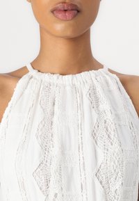 Haut sans manches blanc avec un col froncé et des détails en dentelle complexes, présentant des motifs géométriques et un tissu doux et texturé.
