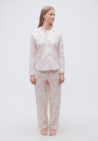 Γυναίκα που στέκεται ξυπόλυτη φορώντας ροζ ανοιχτό ροζ πουκάμισο πιτζάμα με λουλουδάτο σχέδιο και αντίστοιχο φαρδύ παντελόνι, κοιτάζοντας προς τα δεξιά της.