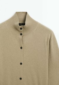 Cardigan beige en tricot avec un col montant côtelé et cinq boutons noirs sur le devant, étalé à plat sur un fond blanc.