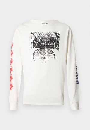 Baltas ilgomis rankovėmis marškinėlis su ryškiu juodu grafiniu piešiniu priekyje ir raudonu tekstu ant kairės rankovės. Audinys atrodo minkštas.