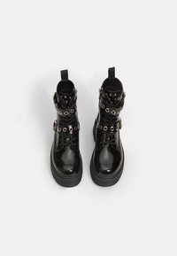 Botas de tobillo de charol negro con suela plana, decoradas con acentos de ojales plateados y correas ajustables. Vista superior mostrada.