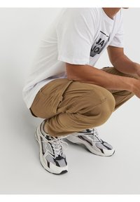 Hellbeige Cargo-Hosen mit mehreren Taschen, kombiniert mit weißen und schwarzen Sneakers, die Mesh- und Gummielemente aufweisen. Weißes T-Shirt mit Grafik.