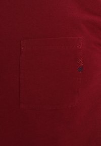 Camiseta de algodón roja con un bolsillo en el pecho y acentos bordados en rojo y azul en el lado, textura suave y diseño ajustado.