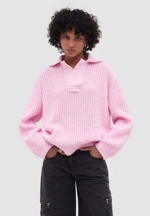 Vrouw met krullend haar, gekleed in een roze geribbelde trui met kraag en zwarte cargobroek, handen in de zakken, tegen een effen grijze achtergrond.