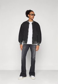 Μαύρο μπουφάν bomber από τζιν με ριμπ μανσέτες, λευκό μπλουζάκι, σκούρο τζιν με καμπάνα, και λευκά αθλητικά παπούτσια, αξεσουάρ με σκούρα γυαλιά ηλίου.