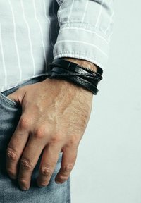 Braccialetti di pelle nera impilati su un polso, caratterizzati da una combinazione di design intrecciati e piatti, con una finitura testurizzata su una camicia chiara.