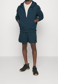 Man wearing dark blue zip-up hoodie and matching shorts, white t-shirt, and black sneakers with gum soles, standing with hands in pockets.