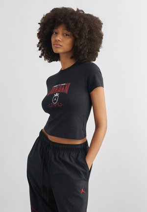 Joven con cabello rizado lleva una camiseta corta negra de Jordan y pantalones deportivos negros con logo rojo, de pie con las manos en los bolsillos.