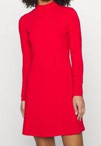 Robe longue rouge à manches longues côtelées, avec un col montant et une jupe évasée. Le tissu a une texture lisse, et la coupe est ajustée à la taille.