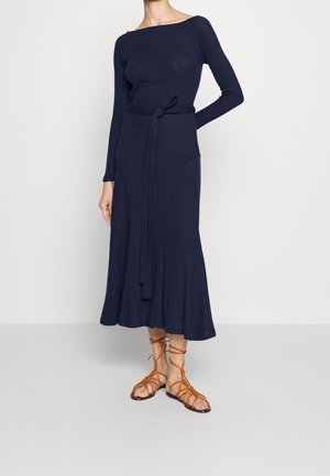 Femme portant une robe midi manches longues bleu marine avec ceinture à la taille et sandales plates marron à lacets sur un fond clair uni.