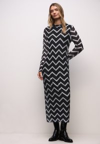 Dlouhé šaty s černobílým vzorem cikcak z průhledného materiálu, s dlouhými rukávy a rovnou siluetou, doplněné černými botami.