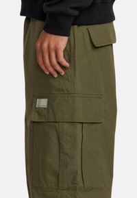 Olive zielone spodnie cargo z fakturowanej tkaniny, wieloma bocznymi kieszeniami oraz naszywką z logo na przedniej kieszeni.