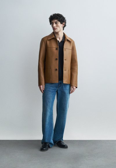 Giovane uomo con capelli ricci indossa una giacca marrone con bottoni, camicia nera, jeans blu e scarpe nere, in piedi davanti a un muro grigio uniforme.