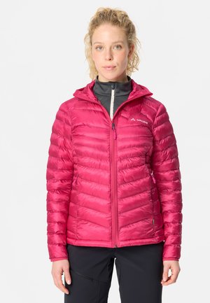 Vrouw met krullend haar die een felroze pufferjack met rits draagt, zwarte broek en een zwarte ritslaag eronder, staand tegen een witte achtergrond.