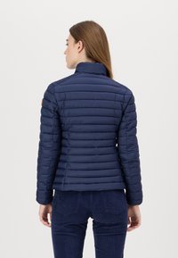 Marine blauwe pufferjas met een hoge kraag, gewatteerd ontwerp en slanke pasvorm. Bevat een oranje logo patch op de linkerschouder. Gladde textuur.