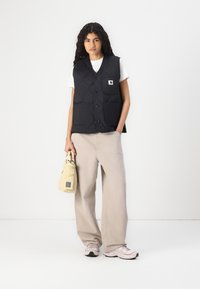 Schwarze gesteppte Weste mit einem Logopatch, zwei Vordertaschen, getragen über einem weißen Hemd, kombiniert mit beigen, weit geschnittenen Hosen und einer kleinen beigen Tasche.