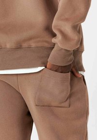 Sudadera marrón con puños y dobladillo acanalados, combinada con pantalones marrones a juego que cuentan con un bolsillo lateral. Tejido suave y texturizado.