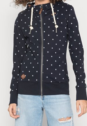 Person wearing a navy blue zip-up hoodie with white polka dots and rope drawstrings, paired with light blue ripped jeans.