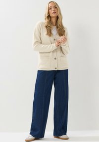Cardigan beige, morbido, con grandi bottoni e due tasche frontali, abbinato a pantaloni in denim blu scuro a vestibilità ampia e sneaker bianche.