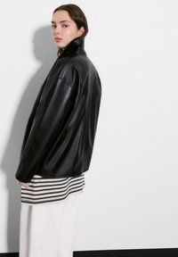 Czarna skórzana kurtka z wysokim kołnierzem, o gładkiej fakturze i dopasowanym kroju, noszona na paskowaną bluzkę i jasne spodnie.