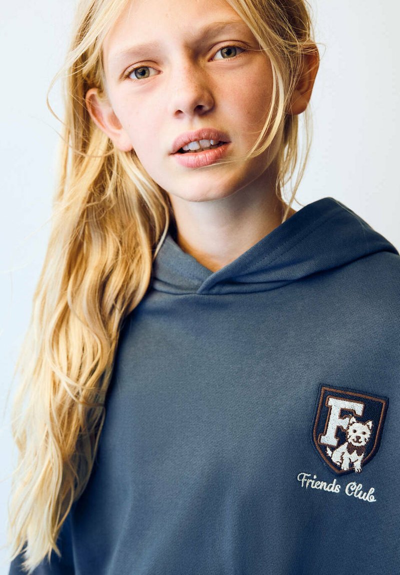 Fille blonde aux longs cheveux portant un sweat à capuche bleu avec un écusson "Friends Club" représentant un chien sur la poitrine, regardant vers la caméra.