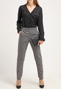 Camicia nera a maniche lunghe con chiusura a bottoni e due tasche sul petto, abbinata a pantaloni grigi a gamba stretta e tacchi neri. Borsa a mano.
