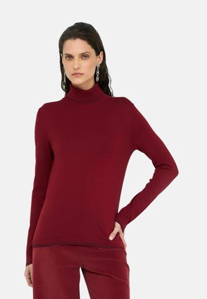 Donna che indossa un maglione aderente a collo alto bordeaux a maniche lunghe e pantaloni abbinati con orecchini a catena metallici, in piedi su uno sfondo semplice.