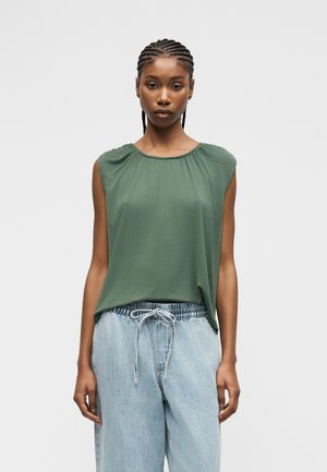 Femme aux cheveux tressés portant un haut vert sans manches rentré dans un jean bleu clair à cordon, debout devant un fond uni.