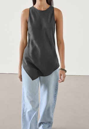 Kvinde iført mørkegrå top uden ærmer med asymmetrisk kant, lyseblå jeans med brede ben og armbånd på venstre håndled.