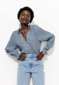 Blouse bleue texturée avec un col à volants et des manches longues, associée à un jean en denim bleu clair taille haute. Détail de bouton remarquable à l'avant.