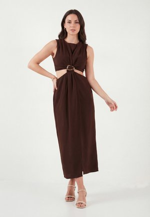 Nő, aki ujjatlan, sötétbarna midi ruhát visel elöl gyűrűs díszítéssel és oldalsó kivágásokkal, bézs nyitott orrú magassarkúval párosítva.