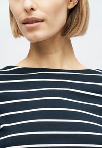 Ženska s kratkimi blond lasmi, oblečena v temno modro majico z belimi vodoravnimi črtami, prikazana od vratu do zgornjega dela prsnega koša.