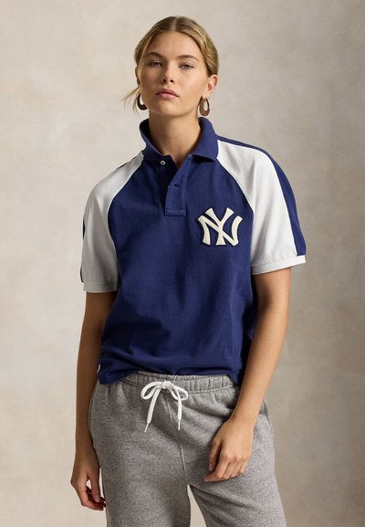 Naine, kes kannab mereväe- ja valget New York Yankees polosärki ning halli dressipükse, seisab ühtlase beežika tausta ees.