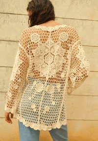 Sweter kardigan na szydełku w kolorze off-white z kwiecistymi wzorami, o otwartym splotcie, z falbanką u dołu i długimi rękawami, w połączeniu z jasnoniebieskimi dżinsami.