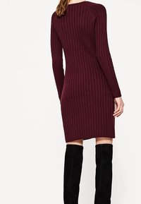 Neule burgundin värinen mekko, jossa on uritettu pinta, pitkät hihat ja tiukka siluetti, yhdistettynä polven yli ulottuviin mustiin saappaisiin.