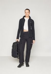 Must must jacket lukuga, valge lühikeste varrukatega särk, mustad cargo püksid ja mustad saapad. Hoides musta seljakotti.